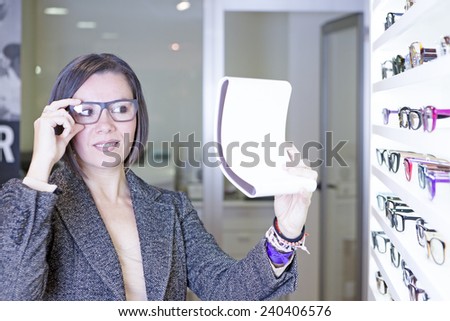 young woman looking in a small mirror is trying on glasses beside a glasses exhibitor at the optical store - focus on the right eye