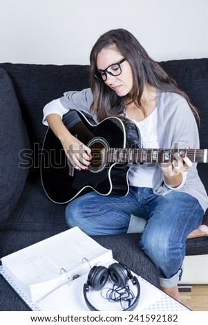 smiling young woman is playing a black acoustic guitar sitting on a sofa at home with a sheet music book and headphones - focus on the girl