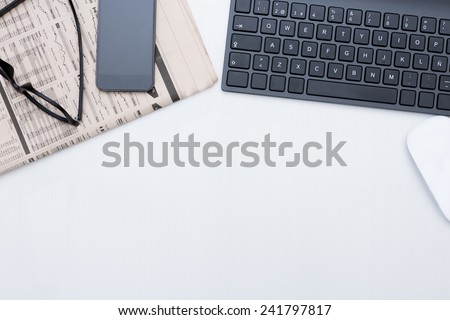 zenithal view of a business desk consisting on a black keyboard, a financial newspaper, a mobile phone, a wireless mouse and a black eyeglasses on a grey desk background - suitable for copy space