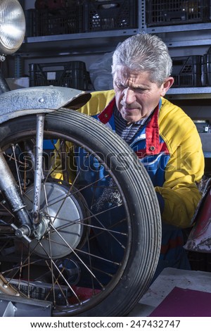 middle aged mechanic sanding the wheel spokes of a classic motorbike with a metal scourer in process of restoration at his workshop - focus on the man face