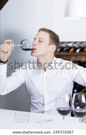 young man on a wine tasting session on the gustatory phase tasting the red wine is writing down in a wine tasting sheet at a restaurant male winemaker smelling a red wineglass - focus on the man face