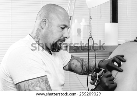 tattooer is tattooing the back of a young woman in the tattoo cabin at his tattoo shop - focus on the man face
