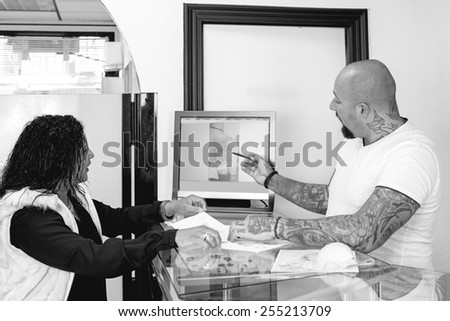 male tattoo artist is showing the drawings in a computer to a young female customer before the tattoo session at the tattoo shop - focus on the man face