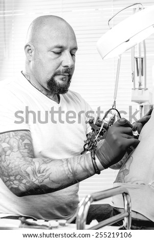 male tattooer is tattooing the back of a young woman in the tattoo cabin at his tattoo shop - focus on the man face