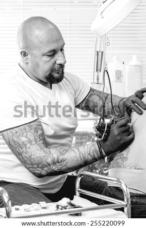 young male tattoo artist is tattooing the back of a young woman in the tattoo cabin at his tattoo shop - focus on the man face
