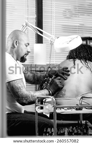 young male tattooer is tattooing the back of a young woman in the tattoo cabin at his tattoo shop - focus on the man face