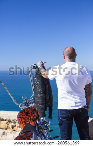 rear view of a chopper motorcyclist standing beside his customized motorcycle with the hand on the handlebar enjoying the view at sunrise - focus on the head