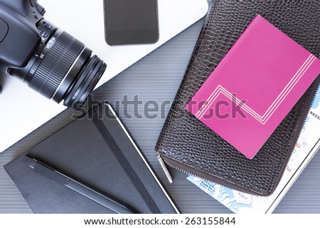 top view of a desktop with a travel stuff consisting on a camera, a passport, a documents case, a phone, a map, a pen, a notebook and a laptop on a desk background