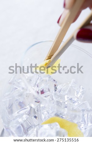 detail of a hand of a woman flavoring inside a gin tonic balloon glass holding a lemon peel with bamboo ice tongs on a flavoring process on a gin tonic preparation session - focus on the lemon peel