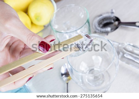 closeup of a woman hand filling a glass with ice from an ice bucket with a bamboo ice tongs on a gin tonic preparation session - focus on the ice cube