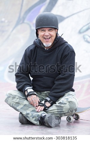 portrait of a smiling old man skater is sitting on his skateboard on a skating park - focus on the face