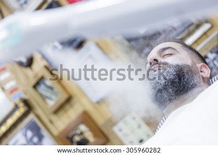face of a customer is being steamed with a steam machine on a beard shaving session in a barber shop - focus on the eye