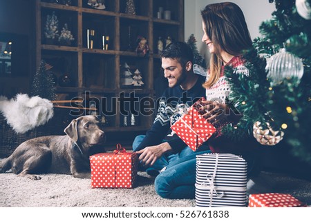 Young laughing couple in sweaters sitting on carpet near fir tree with dog and holding presents.