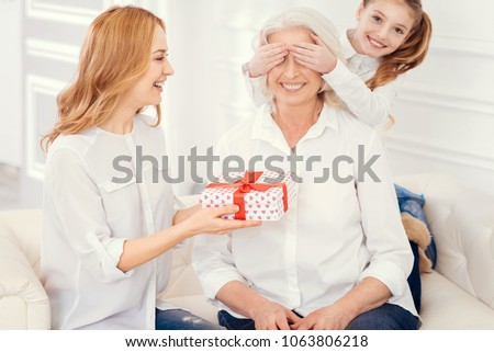 We have a gift for you. Full of love little girl covering up eyes of her cheerful grandmother while a radiant mother giving a beautifully wrapped present and congratulating the granny.
