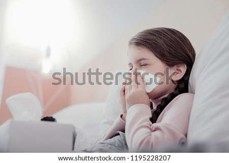 Unpleasant condition. Close up of small pale girl using napkins while keeping her eyes closed and sneezing