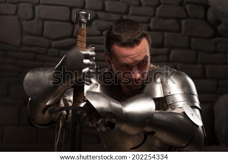Closeup portrait of medieval knight looking down and kneeling with sword on a dark stonewall background