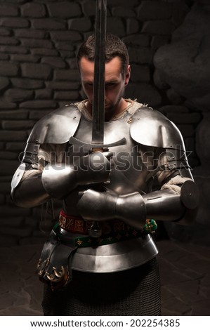 Waistup portrait of medieval knight keeping sword on chest on a dark stonewall background