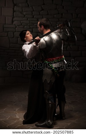 Beautiful couple in historical costumes, medieval knight with sword and lady hugging and petting on dark stonewall background