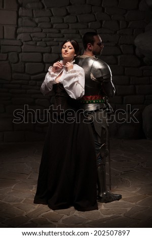 Beautiful couple in historical costumes, medieval knight with sword and lady posing back to back on dark stonewall background