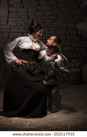 Full length portrait of couple in historical costumes, medieval knight giving rose to beautiful brunette lady sitting on his knee, on dark background