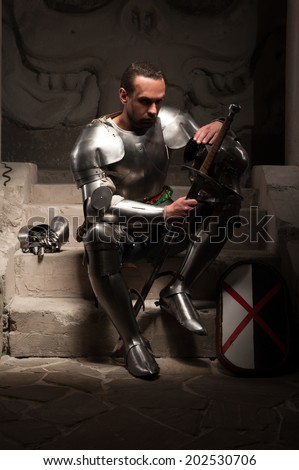 Portrait of medieval knight in armor with shield and sword sitting on steps of ancient temple with skull, dark background