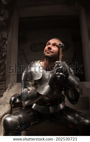 Waist up portrait of smiling emotional knight with sword looking up, on dark stone wall background