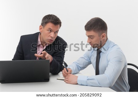 Senior and junior handsome business people discuss something sitting at desk, employee writing in notebook, chief working on laptop, isolated on white