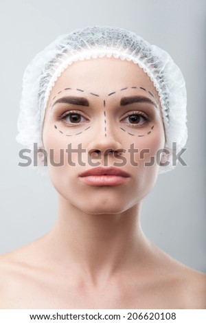 Beautiful young woman in surgical hat with perforation lines on her face looking at camera, before plastic surgery operation, isolated on grey