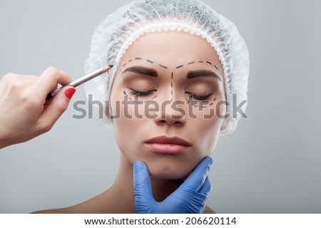 Beautiful young woman with her eyes shut, in surgical cap with perforation lines on her face before plastic surgery operation. Beautician drawing lines on face. Isolated on grey