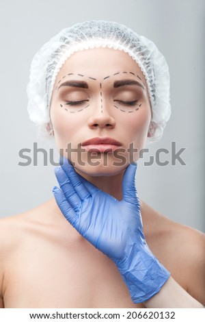 Beautiful young woman with her eyes shut, in surgical cap with perforation lines on her face before plastic surgery operation. Beautician touching face. Isolated on grey