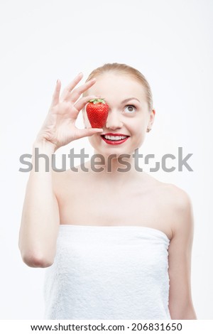 Waist up portrait of a young attractive smiling woman having fun with strawberry, covering her eye, isolated on white