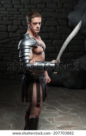 Waist up side view portrait of young attractive warrior gladiator with muscular body posing with sword on dark background. Concept of masculine power, strength