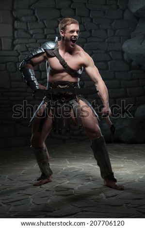 Full length portrait of young attractive warrior gladiator with muscular body posing with two swords, screaming on dark background. Concept of masculine power, strength
