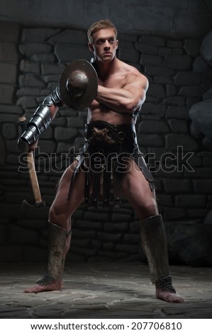Full length portrait of young attractive warrior gladiator with muscular body holding shield and axe, posing on dark background. Concept of masculine power, strength