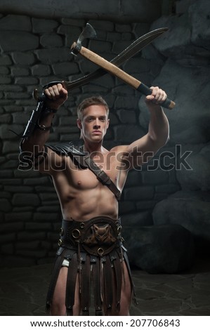 Half length portrait of young attractive warrior gladiator with muscular body holding sword and axe on dark background. Concept of masculine aggressive power, strength