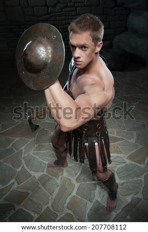 Top view portrait of young attractive warrior gladiator with muscular body holding shield and axe, defending on dark background. Concept of masculine power, strength