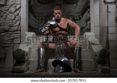 Full length portrait, gladiator in armour sitting on steps of ancient temple with helmet and sword, on dark background. Concept of masculine power, strength