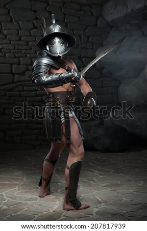 Full length portrait of warrior gladiator with muscular body in helmet holding sword, sideview on dark background. Concept of masculine power, strength