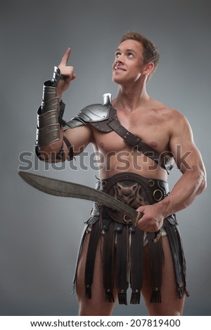 Half length portrait of young handsome muscular man gladiator in armour pointing up holding sword isolated over grey background