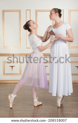 Two young beautiful ballet dancers posing with chocolate, one dancer keeps other from eating a bar of chocolate, full-length portrait in classical interior. Concept of diet or wellness