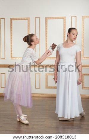 Two young beautiful ballet dancers posing with chocolate, one dancer offering a bar of chocolate to another, full-length portrait in classical interior. Concept of diet and wellness