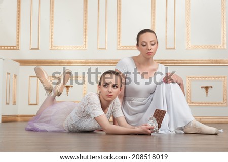 Two young beautiful ballet dancers lying on the floor with a bar of chocolate in classical interior. Concept of diet, wellness
