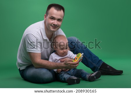Happy handsome Caucasian father sitting on the floor with his son baby boy playing with toy cellphone, isolated on green