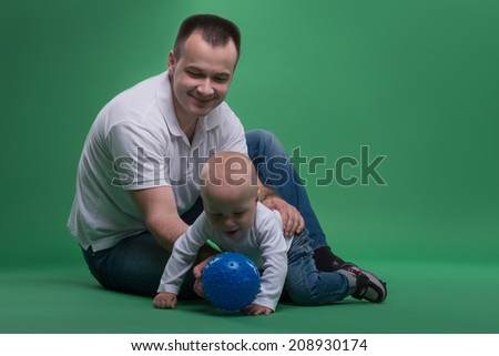 Happy handsome Caucasian father and his son baby boy sitting on the floor and playing with a blue ball, isolated on green