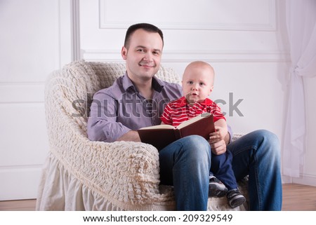Portrait of young handsome smiling father sitting in cozy armchair holding his toddler son on knees and reading a book, interior shot