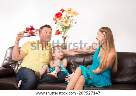 Happy family portrait, little cute daughter sitting on a sofa with beautiful blonde mom and handsome father showing a present, family concept