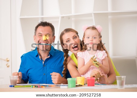 Family portrait, happy caucasian father, beautiful mother and daughter posing while painting a picture with watercolours, handsome father holding a paintbrush in the mouth, interior shot