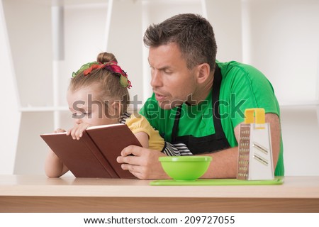 Half length portrait of happy funny handsome man and his cute little daughter wearing apron and preparing to cook with cooking book, grater and bowl, studio shot
