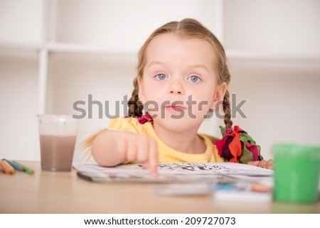 Closeup portrait of little beautiful kid, cute girl painting and pointing at her drawing with finger, pens and paints on the desk, interior shot