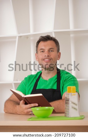 Half length portrait of happy funny handsome man playing fool in the kitchen, wearing apron and preparing to cook with cooking book, grater and bowl